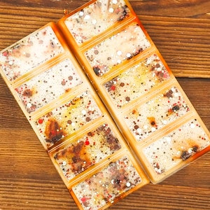 May include: Two rectangular wax melt bars with a creamy white base, speckled with red, brown, and silver glitter. The bars are divided into six sections and have an orange border. The background is a wooden surface.