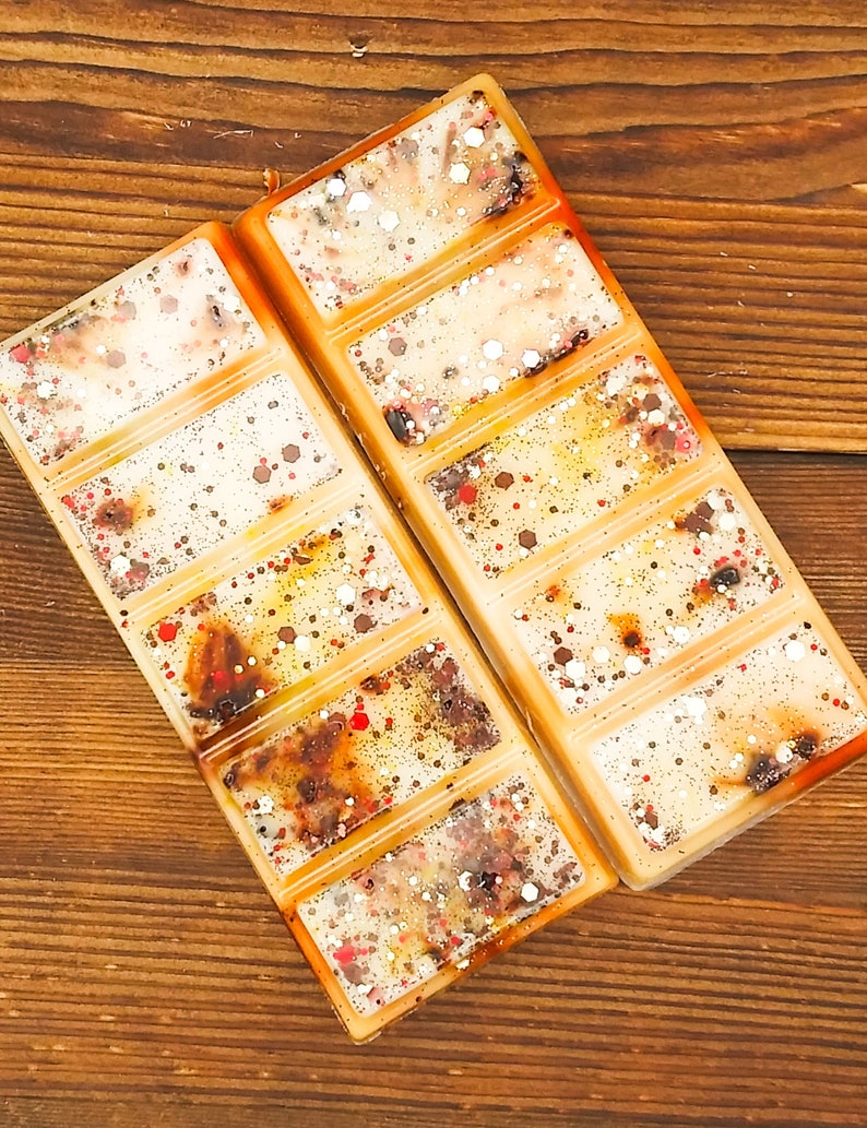 May include: Two rectangular wax melt bars with a creamy white base, speckled with red, brown, and silver glitter. The bars are divided into six sections and have an orange border. The background is a wooden surface.