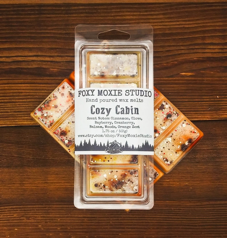 May include: A clear plastic clamshell package containing hand-poured wax melts. The label reads "Cozy Cabin" with scent notes. The wax melts are a light orange colour with flecks of red, brown, and white. The package contains 50g of wax melts.
