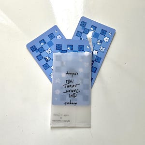 May include: A light blue tarot card set with a floral and checkered design. The set includes three cards and a translucent sleeve with handwritten text that reads "droger's MINI TAROT".