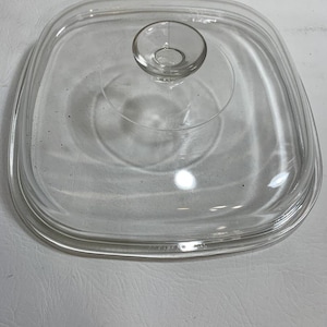 May include: A clear, square Pyrex lid with a rounded handle. The lid is made of transparent glass and has a slightly raised edge. The handle is circular and has a small hole in the center. The lid is designed for cookware.