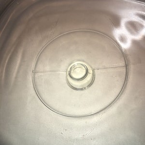May include: A clear, glass lid with a circular design and a central knob. The lid has a slightly curved shape and appears to be for a casserole dish or similar cookware. The glass is transparent, allowing a view of the contents below.