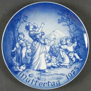 May include: A blue and white porcelain plate with a scene of a mother and her children. The mother is holding a baby in her arms, and the other children are playing around her. The plate is decorated with a blue and white floral pattern and the text "Muttertag 1975".
