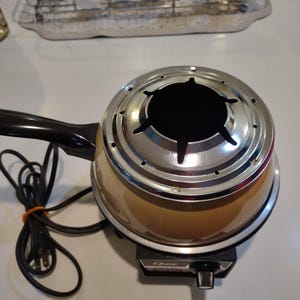 May include: A vintage yellow and chrome electric fondue pot with a black handle and a cord. The pot has a round base with a knob on the front and the word "Oster" printed on it.