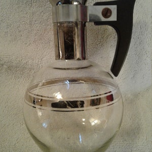 May include: A clear glass pitcher with a silver-colored top and a black plastic handle. The pitcher has a silver band around the middle.