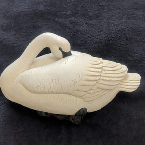 May include: A cream-colored swan figurine with a curved neck and detailed wing feathers. The swan has a black beak and is resting on a dark base. The sculpture is made of a solid material and has a textured surface.