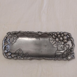 May include: A rectangular pewter-colored tray with a grape vine design. The tray's border features detailed grape clusters and leaves. The center of the tray is smooth and empty, providing space for serving or display.