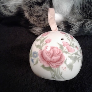 May include: A white ceramic ornament with a floral design featuring pink roses, green leaves, and blue flowers. The ornament has a hole at the top for hanging.
