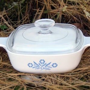 May include: White casserole dish with a glass lid and blue floral design. The dish has a square shape with rounded corners and two handles.