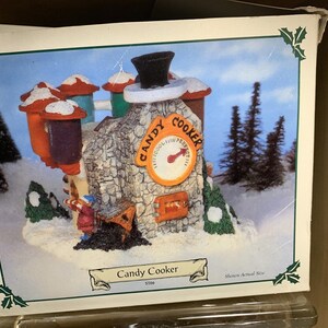 May include: A miniature ceramic Candy Cooker building with a red roof, a chimney, and a thermometer that reads "Candy Cooker Perfect". The building is surrounded by snow and small trees.
