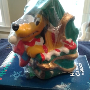 Puede incluir: Una vela de Navidad amarilla y verde con un perro de dibujos animados que sostiene una media. La vela está envuelta en plástico y se encuentra sobre una caja azul con el texto "Holiday Cannons by Enesco".