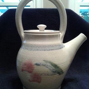 May include: A white ceramic teapot with a rounded body, a large handle, and a lid with a knob. The teapot is decorated with a floral design in shades of pink, green, and blue.