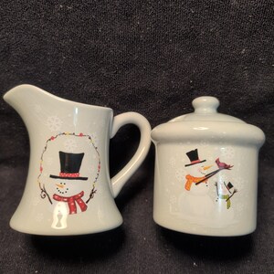 May include: A light gray ceramic creamer and a matching lidded sugar bowl. The creamer features a snowman in a black top hat and red scarf inside a string of lights. The sugar bowl shows two snowmen, one with a red cardinal on its arm.