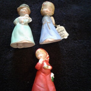 May include: Three porcelain figurines of girls dressed in holiday attire. The girl on the left is wearing a light blue dress and holding a white dove. The girl in the middle is wearing a light blue dress with gold trim. The girl on the right is wearing a red dress with a white collar and a green scarf.