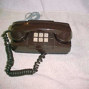 May include: A brown rotary dial phone with a coiled cord. The phone has a keypad with 12 buttons and a rotary dial.