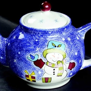 May include: A blue and white ceramic teapot with a snowman design. The teapot has a lid with a red knob and a snowflake pattern. The snowman is wearing a green hat, a red scarf, and a green scarf. The teapot is decorated with a blue and white snowflake pattern.