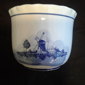 Puede incluir: Una maceta de cerámica blanca con una escena de molino de viento azul y blanco. La maceta tiene un borde festoneado y un diseño azul y blanco.