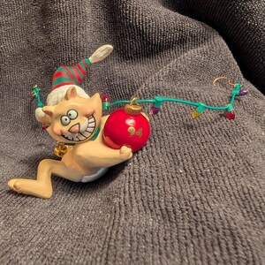May include: A ceramic Christmas ornament of a cat wearing a Santa hat and holding a red ornament. The cat is wearing a green and red striped stocking and has a string of lights wrapped around it.