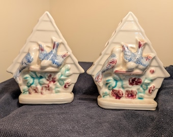 May include: Two ceramic birdhouse-shaped salt and pepper shakers. Each shaker is white with a blue bird and floral design. The birds have red accents. The shakers are on a dark blue surface.
