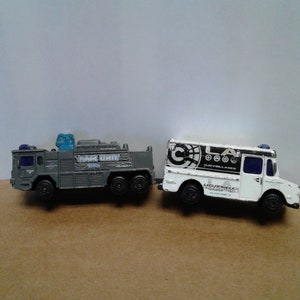 May include: Two toy trucks, one gray with "SWAT UNIT" printed on the side and the other white with "LOS ANGELES SURVEILLANCE" printed on the side.