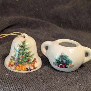 May include: Two small white ceramic Christmas ornaments. One is a bell with a green Christmas tree and presents. The other is a round ornament with a green Christmas tree and presents.