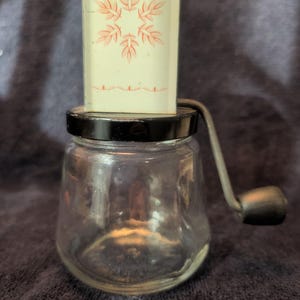 May include: A clear glass jar with a black lid and a hand crank mechanism. The top of the lid features a rectangular cream-colored panel decorated with a pink leaf-like snowflake pattern and a thin pink horizontal line near the bottom edge.