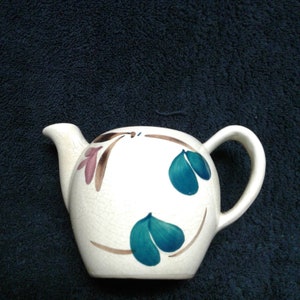 May include: A white ceramic teapot with a brown and pink floral design and blue leaves. The teapot has a round body and a handle.