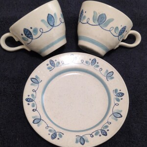 May include: A set of vintage ceramic teacups and saucer. The cups and saucer are white with blue floral designs and a blue stripe. The teacups have handles. The saucer has a matching blue floral border.