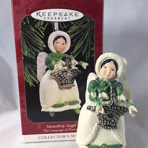 May include: A white porcelain angel ornament with green wings and a green dress. The angel is holding a silver basket filled with white snowdrop flowers. The ornament is part of a Hallmark Keepsake Ornament collection and is titled "Snowdrop Angel The Language of Flowers Collector's Series".