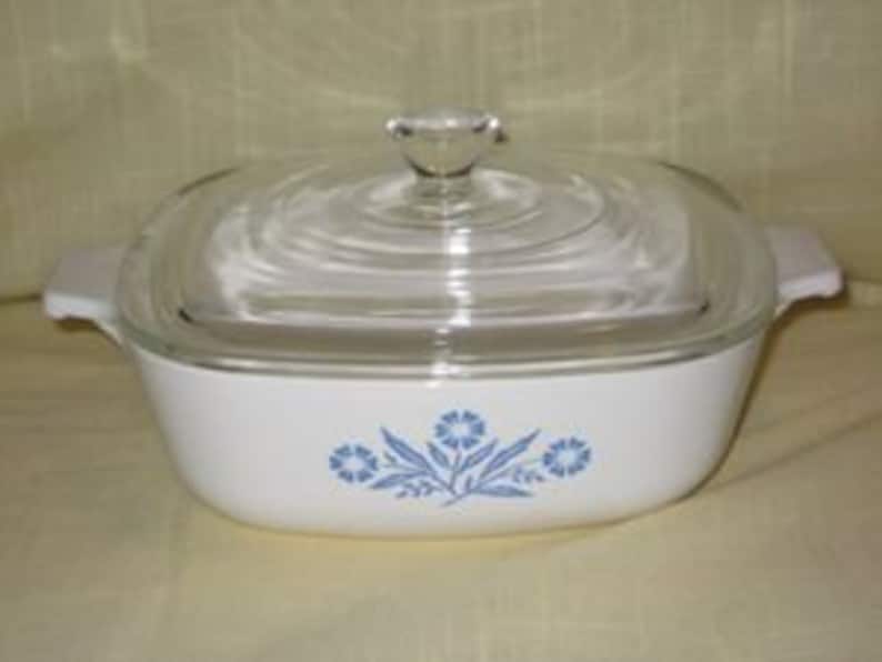 May include: A white square baking dish with a blue floral design and a clear glass lid. The dish has two handles on the sides.