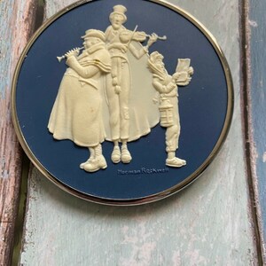 May include: A round, blue and white ceramic plate with a raised image of four carolers. The carolers are dressed in white and are playing musical instruments. The plate is framed with a silver rim and has the text "Norman Rockwell" at the bottom.