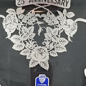 May include: A silver tray with a black background and a silver design of roses and leaves. The tray has a silver banner that says "25th Anniversary". A blue label with the text "Sterling Silver Non-Tarnish" is attached to the tray.