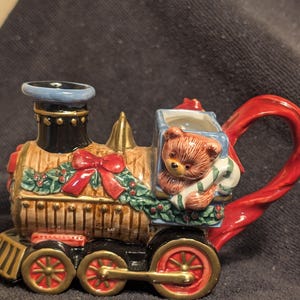 May include: A ceramic teapot shaped like a Christmas train with a teddy bear holding a candy cane in the caboose. The train is decorated with red and green holly and a red bow.