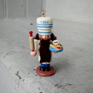May include: A miniature baker ornament with a white hat featuring blue stripes and the year "1994". The ornament has a brown beard, a white coat, and holds a rolling pin and a small pie. It stands on a round, reddish-brown base.