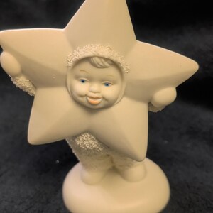 May include: A cream-colored figurine of a child with a smooth face, blue eyes, and orange lips, framed by a large five-pointed star. The figure has a textured, snow-like body and stands on a smooth, round base against a dark background.