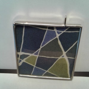 May include: A silver-framed pendant with a geometric pattern of blue, green, and white lines on a white background.