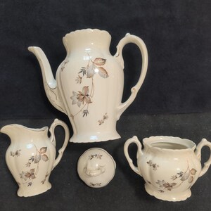 May include: A cream-colored porcelain tea set consisting of a teapot, a creamer, a sugar bowl, and a separate lid. Each piece features a delicate floral pattern with brown and grey leaves and gold trim along the scalloped edges and handles.