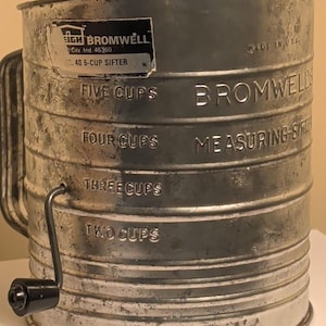 May include: Vintage metal sifter with a handle and a black knob. The sifter has embossed markings for measuring, including "FIVE CUPS" and "FOUR CUPS." The label reads "Leigh Bromwell" and "40 5-CUP SIFTER." Made in the USA.