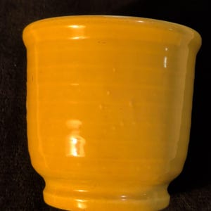 May include: A small, yellow ceramic cup with a rounded top and a slightly flared base. The cup has a smooth, glossy finish and appears to be handmade. The color is a vibrant, sunny yellow.