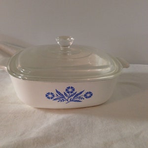 May include: White casserole dish with a glass lid and a blue floral design. The design features three blue flowers with green leaves.