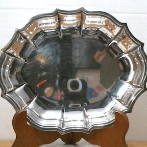 May include: A silver serving dish with a scalloped edge and a shiny finish. The dish is on a wooden stand.