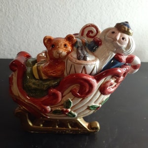 May include: A ceramic Christmas sleigh figurine with a Santa Claus figure, a teddy bear, and a drum. The sleigh is decorated with holly and has a gold base.