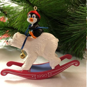 May include: A white polar bear rocking on a red and blue rocking horse with a penguin wearing a red hat riding on its back. The rocking horse has a gold bell hanging from it and the year 1990 is printed on the base.