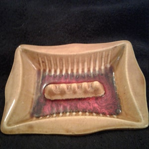 May include: A brown ceramic ashtray with a red and white glaze. The ashtray has a rectangular shape with rounded corners and a raised center section for cigarettes.