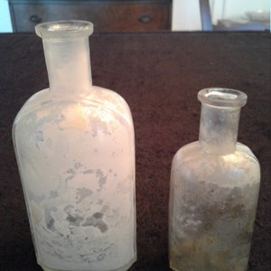 May include: Two clear glass bottles with frosted glass and a textured surface. The bottles are different sizes and shapes.
