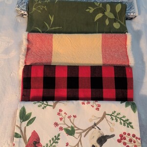 May include: Five folded fabric items are stacked. The top item is a blue and white paisley pattern. Below it is a green fabric with a leaf design. Next is a red and yellow plaid. A red and black buffalo check is below that. The bottom item has a bird and berry design.