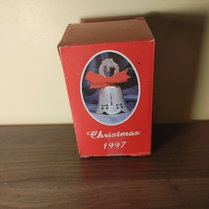 May include: A red box with a white porcelain bell with a red ribbon and a wreath on top. The box says "Christmas 1997".