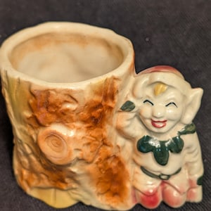 May include: A ceramic planter shaped like a tree stump with a smiling elf figure leaning against it. The planter is painted in shades of brown, green, and yellow.