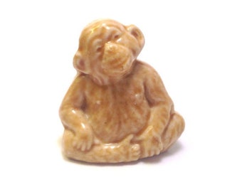 May include: A small ceramic figurine of a brown monkey sitting with its legs crossed. The monkey has a detailed face and fur.