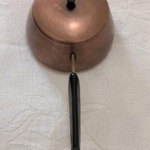May include: A copper-colored, bell-shaped container with a black handle and a small black knob on top. The handle is long and rectangular. The container has a shiny, reflective surface.